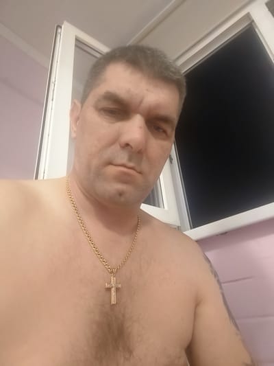 A shirtless man with short, graying hair and a serious expression is positioned in the foreground, looking directly at the camera. He is wearing a gold chain with a cross pendant. The background features a partially opened window with white trim and a dark pane, suggesting it might be nighttime or a room with drawn blinds. The walls appear to be a light pink color, and there's a white ceiling visible above. The man's torso and shoulder are visible, showing light body hair and a tattoo on his left shoulder. The overall scene suggests an indoor setting, possibly a home. No specific location in Petrovsk, Russia, can be determined from the visual information provided.