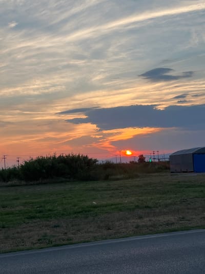 The image depicts a rural landscape at sunset, with a bright orange sun partially obscured by clouds on the horizon. The sky is a gradient of soft oranges, yellows, and blues, with wispy clouds stretching across. In the foreground, there is a grassy field bordered by a paved road. To the left, a dense line of green shrubbery runs along the edge of the field, with several utility poles visible behind it. To the right, a large, multi-colored structure, possibly a building or storage unit, is situated. The overall scene is tranquil and serene, capturing the beauty of a natural sunset. There are no people visible in the image. The location is identified as Chaidefto, Greece.