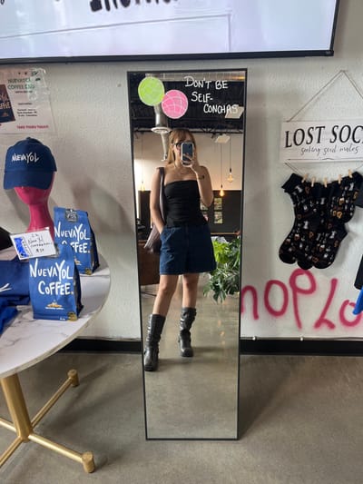The image depicts the interior of Nueva Yol Coffee, located in El Paso, United States. A woman is centered in a full-length mirror, taking a selfie with a smartphone held in her right hand. She wears eyeglasses, a black strapless top, blue denim shorts, and black mid-calf boots, with a dark shoulder bag on her left shoulder. The mirror's surface displays the phrase "DON'T BE SELF-CONCHAS" in white marker, flanked by a green and a pink concha illustration.
To the left of the mirror, a white circular table with a gold base presents merchandise, including multiple blue bags of "NUEVAYOL COFFEE" and a blue "NUEVAYOL" baseball cap on a pink mannequin head. A price tag indicates "1 lb NUEVA YOL COFFEEBAGS $28." A poster detailing "NUEVAYOL COFFEE BAG" information is affixed to the wall above, beneath a large, wall-mounted monitor.
To the right of the mirror, a wooden sign hangs, reading "LOST SOCS seeking soul mates," with several pairs of patterned socks clipped to a string below. The lower section of the white wall to the right features pink spray-painted graffiti forming the word "shop." The establishment's interior features concrete flooring, white textured walls, exposed ceiling light fixtures, and a large green plant partially visible behind the woman's reflection.