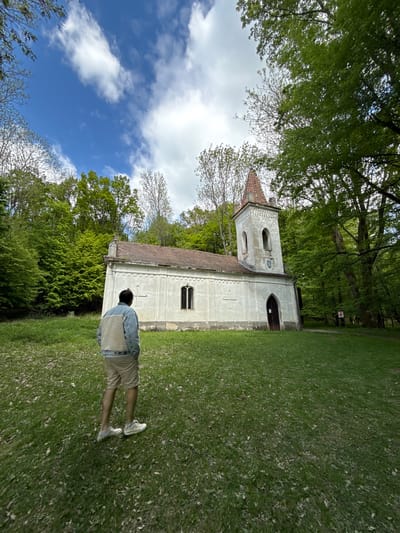 The image shows a person standing on a grassy area, facing a white church-like structure with a tower. The building has a red tile roof and what appear to be arched windows and doorways. The area is surrounded by green trees, and there's a partly cloudy sky visible in the background.
This is likely the Church of St. Emeric in Častá, Slovakia, based on the architectural style and setting. The image captures a scene of someone possibly visiting or observing the church in a natural, outdoor environment.