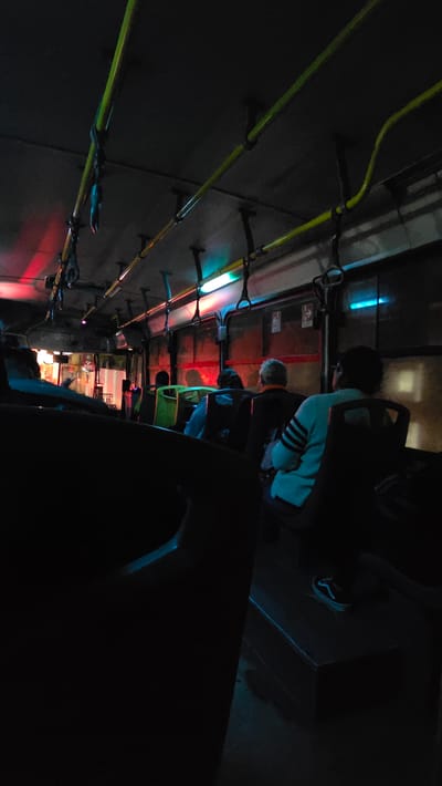 The image depicts the interior of a public transportation vehicle, possibly a bus, in Tlaquepaque, Mexico. Several passengers are visible, including one wearing a light blue sweater with dark stripes and another in a bright green vest. The lighting is dim with artificial colored lighting of red, green, and blue emanating from overhead fixtures. Yellow handrails run along the ceiling of the vehicle. The image is shot from a low angle, making the interior appear elongated. The general environment appears urban and dimly lit outside.