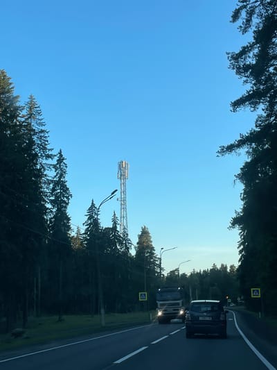 The image captures a road scene in Smolyachkovo, Russia, under a clear blue sky. A tall telecommunications tower is visible in the distance, situated amongst a dense forest of evergreen trees. The road itself is paved with white lane markings and is bordered by the dark silhouettes of trees on both sides. A white truck with its headlights on is driving on the left side of the road, while a dark-colored car is following behind it. Yellow pedestrian crossing signs with a blue pedestrian icon are posted on the right side of the road. The overall environment suggests a rural or semi-rural setting with a road passing through a forested area.