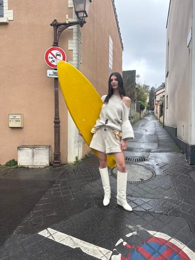 A female subject stands holding a bright yellow surfboard on a wet urban street in Les Lilas, France. The subject, positioned centrally, faces forward with the surfboard held vertically against her right side. She wears a light-colored, off-the-shoulder knit sweater with flared sleeves, light beige high-waisted shorts featuring circular decorative elements on the left side, and white knee-high boots.
The street surface consists of wet cobblestones and asphalt, marked by a foreground 'no parking' symbol (white X over red and blue), and a central manhole cover. To the subject's left, an ornate dark lamppost is attached to a light brown building, displaying a red-bordered 'no motor vehicles' sign above a partially visible white sign reading 'SAINT RIVERAIN'. A utility box and a concrete block are visible at the building's base. The scene recedes into a narrow, overcast alleyway between buildings.