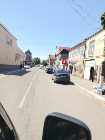 This is a street-level view captured on a sunny day, likely during the daytime. The scene is a street in Kizilyurt, Russia, with buildings lining both sides of the road. Cars are visible on the road, suggesting it is a moderately trafficked area. In the foreground, a reflection of what appears to be a car's side mirror is visible, with the word "ART" faintly reflected on its surface.

On the left side of the street, there are a few buildings with light-colored facades and windows. A satellite dish is mounted on one of these buildings. Further down the street on the left, two people are walking on the sidewalk.

The right side of the street features a row of buildings with storefronts. One building has a prominent sign with the number "2" in a red circle, and other signs with Cyrillic text, including one that reads "ФОТО" (FOTO) and another that appears to be a business name. Further down, there is a sign that reads "КОМПЬЮТЕРЫ" (COMPUTERS). Several trash bins and discarded items are piled on the sidewalk near the buildings.

The sky is clear and blue, indicating good weather. The presence of power lines strung across the sky is also noticeable. The overall impression is that of a typical urban street scene in a Russian town on a bright, clear day.