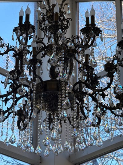 A detailed, low-angle shot captures a chandelier with an elaborate, dark metal frame adorned with numerous clear, sparkling crystals. The chandelier features several candle-style bulbs, emitting a soft light. In the background, through what appears to be a window or skylight, bare tree branches are silhouetted against a pale blue sky. The location is Galevo, Russia.