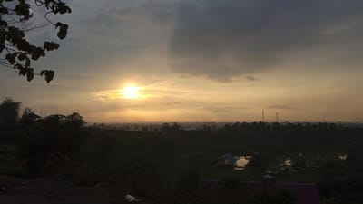 A landscape photograph depicts a scene in Uruekanoe, Indonesia at dusk or dawn. The sun, positioned centrally in the sky, emits a bright glow that is partially obscured by clouds. The sky above the sun is a lighter shade of yellow and fades into darker grays towards the top of the frame.
In the foreground, there are dark silhouettes of trees and foliage. A cluster of leaves is visible on the left side of the frame. In the midground, there's a topographical depression, which may be part of the city. Also, in the midground is a small body of water with a structure built partially over it. The foreground and the midground is mostly dark.
The horizon line contains what appear to be trees and structures, including towers, obscured by the haze. A rooftop of a building can be seen in the bottom right of the scene.
Overall, the scene appears to be a combination of natural landscape and urban development viewed through a lens of diffused light.