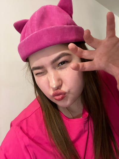 A young woman is posing for a selfie. She is wearing a pink hat with cat ears and a matching pink shirt. She is winking with one eye and has her hand raised in a playful gesture. She is likely located indoors based on the light and wall in the background. There is no evidence to suggest she is in Pridorozhnyi, Russia.