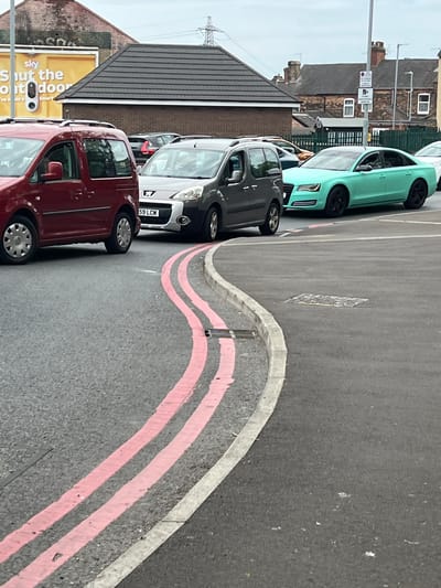 The image displays a row of parked cars on a street in Stoke-on-Trent, United Kingdom. The primary subjects are the vehicles themselves, including a teal Audi A5, a dark grey Citroën C4 Picasso, a lighter grey Fiat Doblo, and a dark red Volkswagen Caddy. The cars are parked alongside a road with distinctive pink and white striped road markings, suggesting a designated parking area or a specific traffic regulation.
In the background, there are buildings with brick facades and pitched roofs, typical of residential or commercial areas in the UK. A prominent electricity pylon stands tall against an overcast sky, indicating the time of day is likely daytime, and the weather appears to be cloudy. A traffic signpost with multiple signs, including one with a purple circle, is visible near the teal car, hinting at parking restrictions or local bylaws.
The scene appears to be a still moment, with the cars stationary and no people clearly visible. The overall impression is one of an ordinary urban environment, capturing a typical streetscape. A large yellow billboard or advertisement for "sky Shut the door" is partially visible on the left side of the image, adding another layer of visual information about the surroundings. The license plate of the Citroën C4 Picasso is readable as "59 LCM".