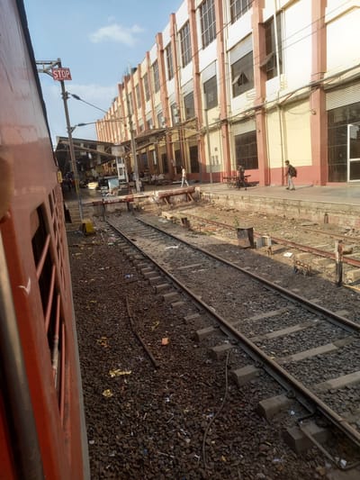 The image depicts a view from inside a train at a railway station in Bhopal, India. The train is stationary next to a platform. Railroad tracks are visible, covered with gravel and scattered debris. A multistory building with light-colored walls and red-painted architectural details runs parallel to the tracks. People are walking on the platform next to the building. A "STOP" sign is visible. Overhead electrical wires are present.