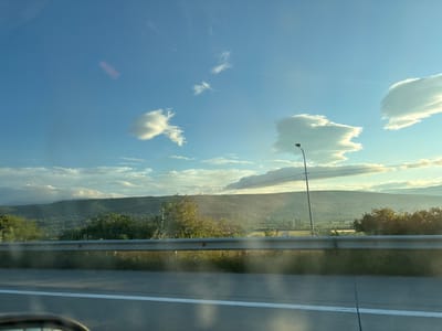 The image shows a view from a vehicle looking out towards a landscape. The sky is blue with scattered clouds, some of which are illuminated by the sun. In the foreground, there is a highway barrier and a road with a white line. Beyond the barrier, rolling hills covered in trees and vegetation are visible, with some buildings scattered throughout. A lamppost is present on the right side of the frame. The overall scene suggests a rural or semi-rural setting during the day, possibly in or near Changilari, Georgia, given the context provided. No people or specific actions are depicted. The lighting indicates it might be late afternoon or early morning.