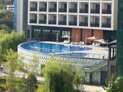 The image shows an outdoor swimming pool area adjacent to a modern hotel building. The hotel features multiple stories with balconies. The pool is kidney-shaped and surrounded by a tiled deck. Several lounge chairs and umbrellas are placed around the pool area, suggesting it is a recreational space. The surroundings include lush green trees and landscaping, indicating an outdoor setting. In the background, a white van is visible on a road near a power line tower. The scene appears to be a resort or hotel complex, potentially located in a suburban or semi-rural area given the greenery and open space. No people are visible in the image. The architecture of the hotel is contemporary, with clean lines and a light-colored facade. A set of outdoor stairs with a glass railing leads down to a ground-level entrance.