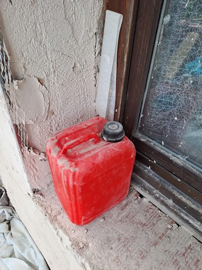 This is a medium shot, eye-level, still photograph of a red jerrycan placed on a rough, off-white textured surface, possibly plaster or concrete. The jerrycan is positioned diagonally across the frame, with its spout facing left. It appears to be dusty and weathered, indicating it might be in a construction or outdoor setting.
Behind the jerrycan, a dark wooden frame runs horizontally, suggesting it is part of a window or door. Beyond this frame, a window pane is visible, obscured by a dense network of white lines, possibly dried paint splatters or frost. White specks are also scattered across the wooden frame and the plastered surface.
The lower part of the frame shows a metal mesh embedded in the plastered surface, with some areas of plaster missing, revealing underlying material. A strip of white material, possibly tape or paper, extends from beneath the spout of the jerrycan towards the left edge of the frame. In the bottom right corner, a crumpled light blue cloth is visible.
The overall scene suggests an unfinished construction site or a neglected area. The lighting appears to be natural daylight, casting soft shadows. There are no people visible in the frame. The location cues of Settimo Torinese, Italy, do not provide further visual information in this image.
There is some text visible on the red jerrycan, although it is partially obscured and difficult to read clearly. It appears to be some form of identification or warning labels, possibly in a European language.