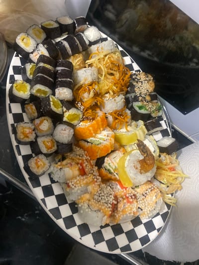 A high-angle, medium shot captures a large platter of assorted sushi rolls arranged on a black and white checkerboard pattern plate. The sushi is diverse, featuring various fillings and toppings. Some rolls are covered in sesame seeds, others have a bright orange roe coating, and some are garnished with crispy shredded ingredients. One roll prominently displays a fried shrimp on top. The setting appears to be indoors, possibly a kitchen or dining area, with a dark countertop visible around the plate and a portion of a black appliance in the background. A roll of white paper towels is also visible in the upper right corner. The lighting suggests it is daytime, but the exact time is not discernible. There is no visible text in the image, and the focus is entirely on the presentation of the food. The sushi itself appears to be freshly prepared, and the variety suggests it is a meal or a substantial appetizer. The overall impression is one of abundance and a visually appealing culinary display.