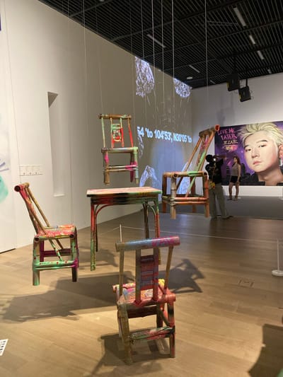 The image shows an indoor art exhibition in Jinniu District, China. In the foreground, a colorful, distressed wooden chair is positioned on a light-brown wooden floor. Behind it, another similar chair is suspended mid-air by thin wires. Further back, a table with a similarly painted surface and a chair hanging above it are visible. To the right, a large painting of a blonde-haired individual with the text "LOVE ME LATER" and Chinese characters is displayed on a wall. Two women are standing in front of this painting. A man is taking a photograph of the artwork. The background features a white wall with a projected image of a celestial body and coordinates. The ceiling has a grid of black beams and lights. The scene suggests a modern art installation with a focus on repurposed and artistically modified furniture.