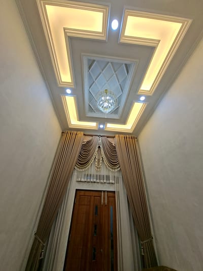 This image captures an interior view of a room, likely a hallway or entrance, in Uleemeuria, Indonesia. The focal point is a sophisticated coffered ceiling with integrated lighting. The ceiling features a central recessed area with a diamond pattern and a decorative chandelier. Surrounding this, a stepped design with warm, recessed yellow lighting forms an angular border. Circular ceiling lights are also visible within this stepped design.
Dominating the lower portion of the image are two tall, wooden doors, likely an entranceway. These doors are flanked by voluminous, taupe-colored curtains that are drawn back to reveal sheer white curtains underneath. The curtains are elaborately draped and adorned with decorative trim and tassels at the top. The walls are painted a neutral, textured light color. The perspective is looking upwards towards the ceiling and the entrance, creating a sense of grandeur and depth. No people are visible in the image. The environment appears to be a well-lit and elegantly decorated indoor space.