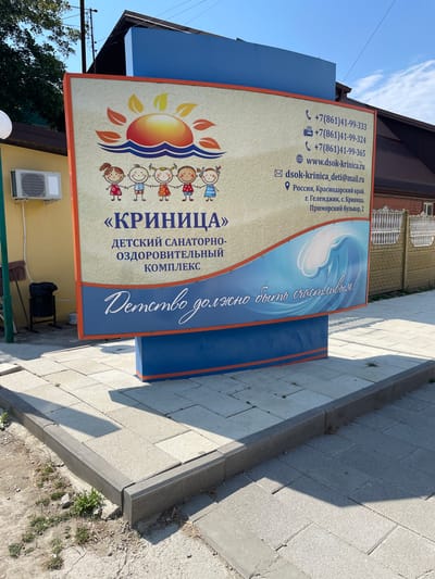 A large, colorful billboard advertising "KRINitsa" Children's Sanatorium and Health Complex stands on a paved sidewalk in Krinitsa, Russia. The billboard features cartoon drawings of children and a sun rising over waves. Contact information, including phone numbers, website, email address, and physical address, is displayed on the right side. The physical address provided is Primorsky Boulevard, 2, Gelendzhik, Krinitsa, Krasnodar Krai, Russia. The billboard is supported by a blue and orange structure. In the background, a yellow building with an air conditioning unit is visible, along with a black trash can and some greenery. The sky is clear and blue, indicating a sunny day.
