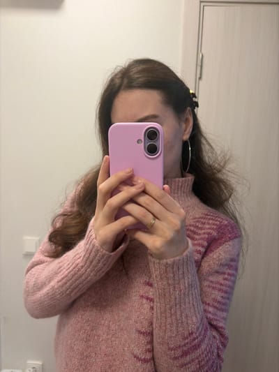 A person holds a light purple phone, possibly taking a selfie. They wear a pink sweater with a textured pattern. The individual has brown hair, styled with a clip and hoop earrings. A simple gold ring adorns their finger. The background features a white wall with a cabinet door visible in the upper left and an electrical outlet in the lower right. The image appears to be taken in a bathroom or similar indoor setting.