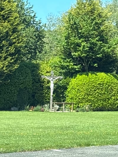 The image shows a religious statue, specifically a crucifix, in a green, natural setting likely a cemetery in Wistaston, United Kingdom. The crucifix is made of wood or stone, with a white or light-colored figure of Jesus attached. The background includes trees, bushes, and a well-maintained lawn. A small metal fence or gate is partially visible near the statue. The scene is shot in daylight, with some shadows indicating the position of the sun.