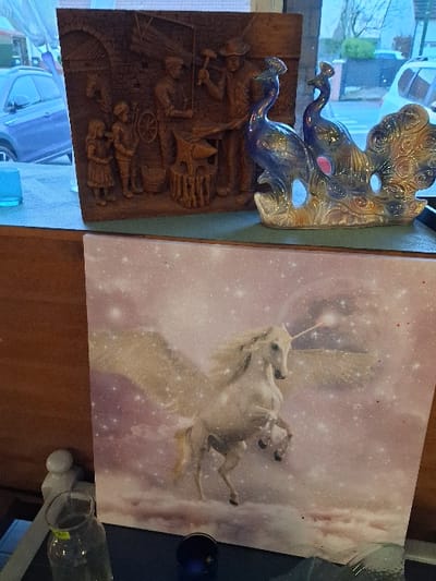 The image shows two mounted artworks. One is a dark-toned relief of workers; above it is a decorative piece shaped like two peacock heads, primarily blue with some gold. The second, larger piece displays a white unicorn leaping against a backdrop of purplish clouds and stars. Several household objects are placed on the furniture surrounding the artwork.