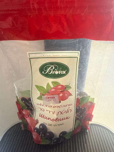 The image shows a clear plastic bag of dried fruit. The bag has a label with the brand name "BioFIX" in an oval green logo at the top. Below the logo, there is an illustration of red berries and dark purple berries with green leaves. There is text in Hebrew on the label. A price tag is visible, showing "₪ 12.90". The background is blurry but appears to be indoors, possibly a kitchen or pantry, with a textured surface below the bag. The location in the city of Dimona, Israel, is not discernible from the image itself; this information is provided contextually.