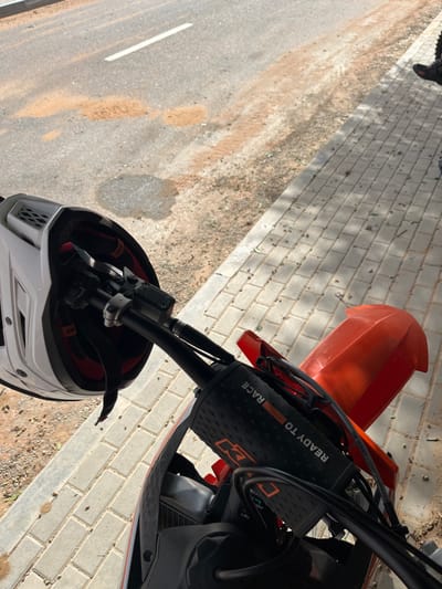 The image depicts a first-person view of a parked KTM dirt bike on a sunny day in Al Madam, United Arab Emirates. A white motocross helmet with red internal padding is suspended from the left handlebar. The handlebar's crossbar pad displays "KTM" and "READY TO RACE" branding. An orange front fender is visible to the right. The motorcycle is positioned alongside a sidewalk composed of rectangular grey paver bricks. To the left of the sidewalk is an asphalt road, characterized by significant accumulations of reddish-brown sand and dirt, with a faint white dashed line marking visible in the distance. Sparse vegetation and debris are scattered on the unpaved ground between the road and sidewalk, and also lightly on the sidewalk itself. In the upper right corner, the lower legs and dark footwear of a person are partially visible, standing on the sidewalk. Strong shadows are cast across the scene, indicating direct sunlight.