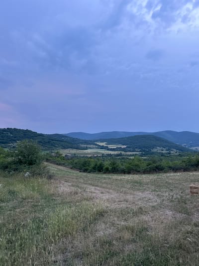 The image depicts a rural landscape in the vicinity of Malka Vereya, Bulgaria, under a twilight sky. The upper approximately one-third of the frame is dominated by a sky displaying hues of muted purple and blue, with scattered, indistinct clouds exhibiting lighter tones. Below the sky, a series of undulating hills and low mountains, densely covered in dark green deciduous foliage, stretch across the middle ground and background. Atmospheric perspective renders the more distant mountains in slightly bluer tones. Below the forested hills, patches of cultivated or fallow land are visible, presenting a mix of lighter green and subdued brown vegetation. The foreground comprises a field characterized by a combination of taller green grasses and drier, shorter brown stubble, indicating either recent harvesting or natural variation in growth. A discernible track or path, marked by less dense vegetation, traverses the foreground field from the lower left towards the center. In the lower right quadrant of the foreground, a single, rectangular hay bale is visible. No people or active interactions are present in the scene. The lighting suggests either dusk or dawn.