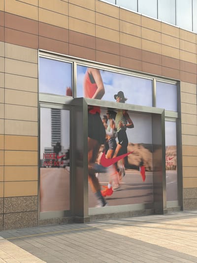 This image captures a section of a modern building facade in Yuhuatai District, China, featuring large advertising displays. The building's exterior is constructed with alternating bands of light beige and brown rectangular panels, arranged in a grid pattern.
Prominently featured are multiple window displays showcasing a Nike advertisement. The main display depicts several people in athletic wear engaged in running. A central figure, a woman wearing a light-colored top, dark shorts, and a cap, is shown mid-stride with other blurred figures around her, conveying a sense of dynamic motion. A large, bold red Nike swoosh logo is superimposed over the runners.
Visible text within the advertisements includes "NIKE" and its iconic swoosh, as well as the slogan "JUST DO IT," both rendered in red against the background imagery of the runners and a bright sky. These elements appear in both the larger central display and smaller adjacent window sections.
The scene is brightly lit, suggesting it was captured during daylight hours. The architectural style is contemporary, characterized by clean lines and mixed materials like glass, metal, and composite panels. The overall impression is that of an urban commercial environment.