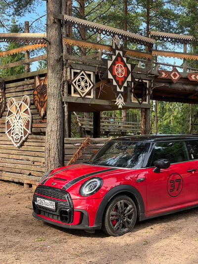 A red Mini Cooper is parked on a dirt road in front of a wooden structure decorated with geometric carvings and animal motifs. The Mini Cooper has black racing stripes on its hood and a black roof. A circular decal with the number "37" is visible on its side. The wooden structure appears to be a gate or entrance, with intricate carvings of animal faces and geometric patterns. The background consists of a forest with tall trees and blue sky. The license plate of the car reads "0267 AA 250" and indicates it is registered in Russia. The scene appears to be located in Ozerki (Krasnodolinskaya), Russia. There are no people visible in the image.