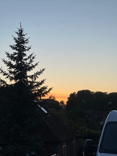 The image captures a scene in High Wycombe, United Kingdom, during twilight. The sky transitions from light blue to a warm orange hue as the sun sets. A vapor trail from an aircraft is visible in the sky. Silhouetted against the sky are the branches of a conifer tree in the foreground, and darker forms that appear to be a combination of trees and buildings in the background, including a house with a dark tiled roof and what appears to be a satellite dish. A partial view of a white van is seen at the top right corner. The scene appears quiet, without obvious human presence or interaction.