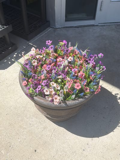A flower-filled planter sits on a concrete surface outside a building. The planter is round and made of a dark, textured material, possibly stone or concrete. It is overflowing with artificial flowers in various shades of pink, purple, and blue, mixed with greenery. In the background, a metal grate is visible, along with dark vertical posts, suggesting an entrance or a covered area. The building facade is light-colored, with a glass window and a door. Sunlight casts a shadow of the planter onto the concrete.