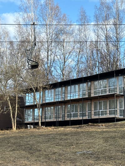 The image depicts the exterior of a multi-story building with numerous windows, likely a residential or commercial structure, situated in what appears to be a wooded area. The location is identified as Galevo, Russia. The building has a dark-colored facade with a grid-like pattern created by the window frames. The windows reflect the surrounding environment, including trees and the sky. In the foreground, there are several bare trees with intricate branch patterns, typical of a winter or early spring scene. A utility pole with a light fixture is also visible among the trees. To the right of the building is a patch of dry grass or vegetation. The sky is blue with some clouds. The overall impression is of a building integrated into a natural setting on a clear day.
