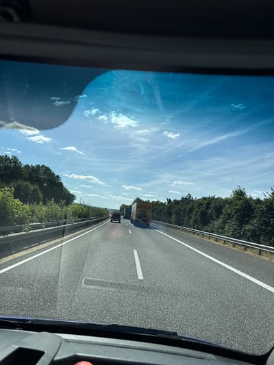 The image shows a view from inside a vehicle traveling on a highway in Laufeld, Germany. The perspective is from the driver's seat, looking through the windshield. The highway is paved with asphalt and has white lane markings. On the right side of the highway, there is a metal guardrail separating the road from a dense forest of green trees. On the left side of the highway, there is also a guardrail and a steeper, wooded embankment.

Several vehicles are visible on the highway. In the foreground, the dashboard of the car is visible, including a dark grey surface and a hint of a red object, possibly a button or a toy. In the middle distance, there is a black SUV driving ahead, followed by a yellow truck with "SPEDLOG" written on its side. Further ahead, other vehicles, including trucks and cars, can be seen on the highway.

The sky is bright blue with scattered white clouds and some wispy cirrus clouds. The sun is shining, casting shadows on the road and highlighting the trees. A wind turbine is visible in the distance, to the left of the highway, amidst the trees. The overall impression is a bright, sunny day on a rural German highway.