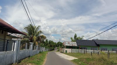 The image depicts a rural street scene in Geulanggang Baro, Indonesia, under a partly cloudy sky. A paved road curves into the distance, lined on both sides by fences and residential buildings. On the left, a house with a red-tiled roof and a light-colored wall is partially visible behind a concrete fence with black metal railings. A tall palm tree stands next to this property. On the right, a long, green building with a dark metal roof is prominent. This building has multiple windows and appears to be a communal structure or school, with a lighter-colored fence in front of it. The area is characterized by lush green vegetation, including numerous trees and grassy verges along the road. Power lines stretch across the upper portion of the image. No people are visible in the frame.