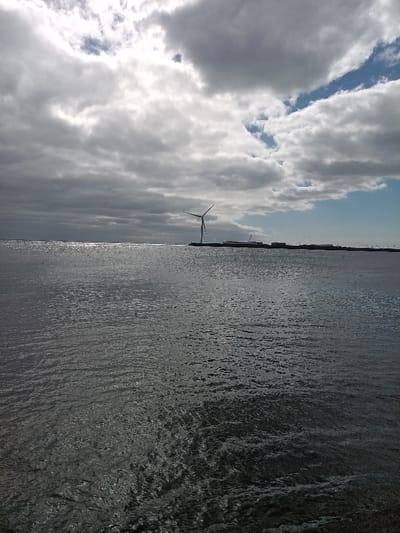 This image depicts a coastal view in Agüimes, Spain, facing a body of water under a partly cloudy sky. The foreground is dominated by the dark, reflective surface of the water, exhibiting subtle ripples and reflections of sunlight across its expanse, particularly a bright streak towards the left horizon. In the midground, a distinct wind turbine with three blades, one appearing partially rotated, stands on a distant landmass or breakwater extending horizontally across the frame. To the right of the wind turbine, various low-lying industrial or port-like structures, including cranes or similar machinery, are visible along the distant coastline. The background features a sky filled with a mixture of dark, heavy clouds and patches of brighter blue sky visible in the upper right quadrant. No people or active interactions are visible in the scene, which appears static.