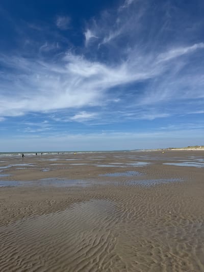 The image depicts a beach in Berck, France. The scene is dominated by a vast expanse of wet sand patterned with ripples, suggesting the recent retreat of the tide. Scattered puddles of water reflect the sky. In the distance, the calm sea meets a hazy horizon. The sky is a bright blue, adorned with wispy white cirrus clouds. Several people are visible as small figures on the beach, some walking and others standing. A few yellow buoys are dotted along the coastline. The overall impression is one of a serene, open beach under a wide sky.
