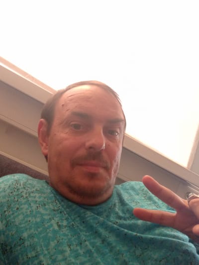 A close-up, waist-up, selfie-style photograph captures a light-skinned man with thinning brown hair, a mustache, and a mole on his forehead. He is wearing a teal t-shirt with a speckled pattern. The man is positioned in the foreground, with his head slightly tilted. His right hand is raised, forming a peace sign, and he is wearing a ring on his index finger. The background is out of focus, but appears to be an interior space with a white ceiling or wall and some beige trim. The lighting is bright, suggesting it might be daytime. There is no indication of the city of Selli, Estonia, in the image itself.