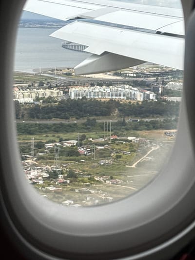 View from an airplane window over Montijo, Portugal. Airplane wing visible in the left of the frame. Landscape features buildings, roads, trees, water, and power lines.