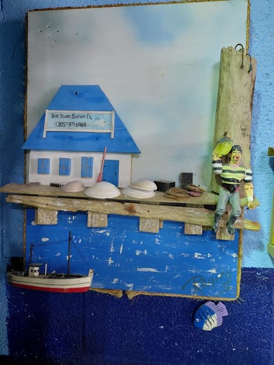 The image shows a handmade artwork of a seaside scene hanging on a blue wall. The artwork is constructed of various materials. It features a small house with a blue roof, labeled "Bone Island Seafood Co. (305) 971-6464." The house appears on a white background, possibly representing the sky or land. A figure resembling a pirate is positioned near what looks like a wooden plank. The scene incorporates shells, pieces of wood, and a small toy boat that looks like it is in the water. A small, stylized fish is attached to the artwork. The background wall has a textured, dark blue section on the right side and a lighter blue section on the left. The scene likely depicts a nautical theme. The Cutler Bay city is only inferred from the area code listed in the artwork.