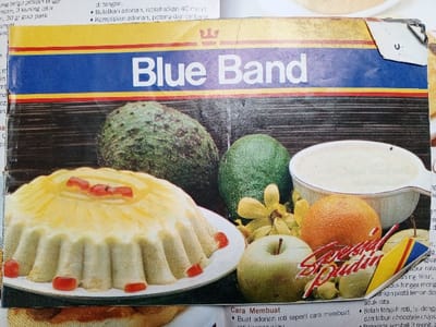 The image shows a recipe book page with a picture of a dessert featuring a Blue Band brand. The dessert appears to be a type of flan or pudding, garnished with red fruit pieces. Avocados, an orange, an apple, and a bowl of a white creamy substance are visible in the background. Part of the recipe is printed in Indonesian, with instructions including steps like "Bulatkan adonan, istirahatkan 40 menit" (Round the dough, rest for 40 minutes) and "Buat adonan roti seperti cara membuat" (Make bread dough like making). The text also references ingredients like flour, egg yolks, and sugar.