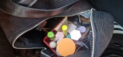 A close-up, top-down view depicts the partially unzipped interior of a dark, likely black, ribbed fabric bag, located in Cenon, France. The bag's zipper is silver-toned. Inside, a red rectangular container holds an assortment of personal items. These include an orange circular cosmetic sponge, two hexagonal sponges (one light pink, one white/light beige), and several cylindrical tubes with colored caps (one green, one yellow, one light purple). A light beige spherical object, a silver-toned eyelash curler, and multiple dark cylindrical objects resembling pens or brushes are also visible within the bag. Various crumpled papers or labels are interspersed among the items. The bag's interior lining is dark. A dark fabric strap is visible across the top, and a portion of a white-striped object is partially visible on the far right side of the bag's exterior. No individuals are present, and no actions are discernible.