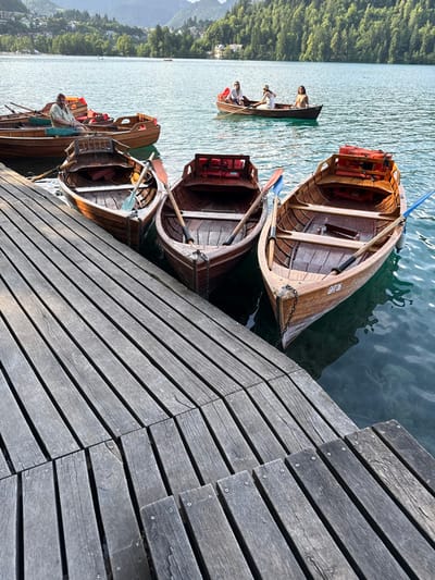 A wooden dock extends into Lake Bled, Slovenia (latitude: 46.364, longitude: 14.091). Three wooden rowing boats are moored to the dock, their bows facing the lake. Two of the boats are empty, with oars resting in the boat. One boat has a man sitting in the back, looking straight ahead. In the distance, another boat is being rowed across the lake by three people, two women and a man. The lake is calm and reflective. Lush greenery surrounds the lake, indicating a forested landscape. The dock is made of rough, weathered planks arranged in parallel rows, and the dock is wet from the lake water.