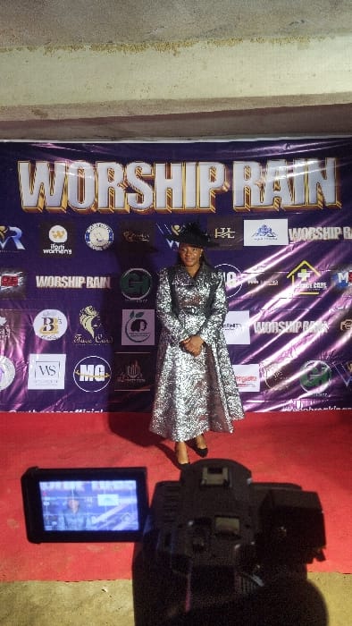 The scene depicts a woman posing for a video recording at an indoor event in Buea, Cameroon. The subject, a woman, stands centrally on a red carpet, facing forward with a slight smile and hands clasped. She is dressed in a long, silver-patterned gown with dark shoulder and waist accents, complemented by a dark, wide-brimmed hat and dark closed-toe shoes. Behind her is a large purple banner displaying "WORSHIP RAIN" in prominent gold and white lettering, surrounded by numerous sponsor logos. In the lower right foreground, a black video camera is partially visible, with its articulating screen displaying a live feed of the woman against the banner, indicating active recording. The upper background reveals a plain, light-colored concrete wall. The specific location within Buea is an event venue associated with the "Worship Rain" event series.
