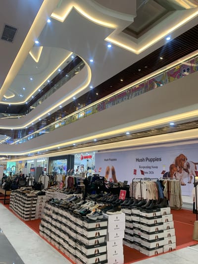 This image captures the bright, multi-level interior of a modern shopping mall, viewed from a lower level looking upwards and across. The architecture features sleek, curved white surfaces illuminated by continuous LED strip lighting and recessed spotlights, creating a sophisticated and clean aesthetic. Dark wood slats are visible on upper ceiling sections, contrasting with the white. Air vents are also visible on the ceiling.

On the right side of the image, appearing as the floor due to the tilted perspective, is a retail area primarily featuring the brand "Hush Puppies." A large banner prominently announces "Hush Puppies Reopening Soon June 2026," suggesting a future event. Below this banner, numerous stacks of Hush Puppies shoe boxes are arranged, along with racks displaying clothing and shoes, including sneakers. A red tag indicating "-10%" and "-20%" discounts is visible on some merchandise.

In the background and on other levels, various other stores and displays can be glimpsed, including a partially visible "TOMMY HILFIGER" sign and a colorful display on an upper railing that seems to read "THE NEW CHAPTER." The overall scene suggests a vibrant commercial environment, likely in a city like Samarinda, Indonesia, given the context. No people are distinctly visible, but the setup implies active retail operations. The bright artificial lighting gives no clear indication of the time of day.