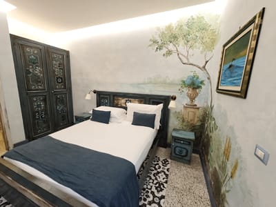This media file depicts a bedroom with a distinctive artistic mural covering the walls. The mural features a serene landscape with a large tree, a body of water, and lush greenery, creating a tranquil ambiance. A framed painting of a duck swimming in blue water hangs on the wall.
The bedroom is furnished with a bed, two bedside tables, and a large, ornate wardrobe. The bed is neatly made with white linens and dark blue accent pillows and a runner. The bedside tables are also painted in a distressed blue style, matching the wardrobe. The wardrobe has intricately carved details and appears to be quite old and grand.
The flooring is a speckled terrazzo style, common in older Italian buildings. A decorative rug with a black and white pattern is placed near the bed. Wall-mounted lamps are positioned above the headboard, casting a warm glow. A light switch is visible on the wall. The lighting in the room is soft and diffused, suggesting it might be evening or a dimly lit day.
The overall impression is one of a charming, perhaps vintage-inspired, bedroom with a strong artistic theme. The setting suggests a private residence or a boutique hotel room in Pozzuoli, Italy, a coastal town known for its history and natural beauty. There is no visible text in the image.