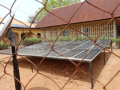 The image shows a solar panel array in Rumde Waziri, Nigeria. The array is made up of several panels arranged in a grid. The panels are mounted on metal frames and are supported by metal poles. The array is located in an open area, surrounded by a chain-link fence. There is a satellite dish mounted on a pole in the foreground. The array is set up in front of a building with a red corrugated tin roof, windows, and a yellow-painted exterior. The building appears to be an administrative or residential structure. There is a low, green bush growing in front of the building. The ground around the array is dusty and covered with dried leaves. The scene is captured from a low angle, through the chain-link fence.