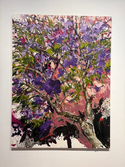 This is a close-up, eye-level shot of an abstract painting displayed on a white wall, possibly in a gallery or art exhibition. The painting is predominantly a vibrant mix of purples, greens, and pinks, with accents of black and white. It depicts an abstracted tree, characterized by thick, gestural brushstrokes and splatters of paint. The upper portion of the canvas is filled with a dense canopy of leaves and flowers rendered in various shades of purple and green, interspersed with pink. The trunk of the tree is a prominent vertical element in the center, painted with lighter tones of white and brown, suggesting texture. The lower part of the painting features darker, more dramatic strokes of black and deep purple, creating a sense of depth and contrast. The overall impression is energetic and expressive. The artwork is framed by the stark white of the wall and the subtle shadow cast by its edge, indicating it is a physical object and not a digital display. The image's context as being located in La Cuesta, Spain is not visually evident from the artwork itself.