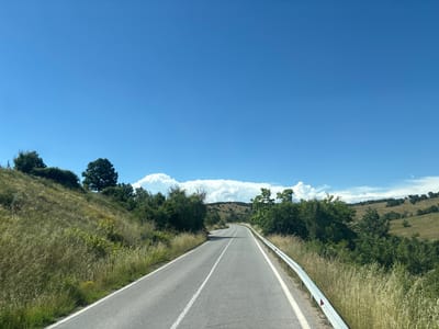 The image shows a two-lane asphalt road winding through a hilly, rural landscape under a clear blue sky with scattered white clouds. The road is bordered by a metal guardrail on the right and a grassy embankment on the left, covered in tall, dry grass and scattered green bushes and trees. In the distance, rolling hills are dotted with trees and fields, suggesting a pastoral setting. The time of day appears to be daytime, likely late morning or early afternoon, given the bright sunlight casting shadows. The weather is sunny and fair. No people or specific objects are prominently featured, focusing instead on the natural scenery and the road as a path through it. The overall impression is one of a scenic drive through the countryside of Mladinovo, Bulgaria, evoking a sense of peace and open space.