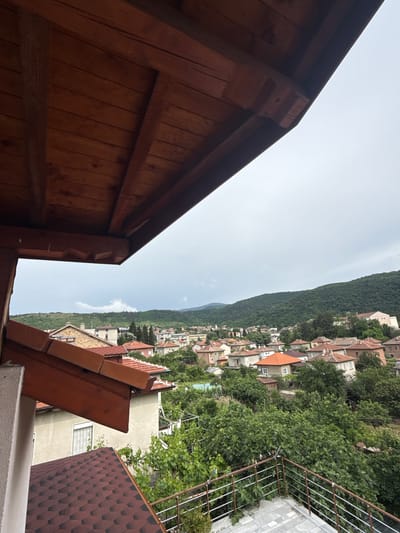 The image, viewed from a high vantage point, captures a panoramic scene of a town nestled in a mountainous landscape under an overcast sky. In the immediate foreground on the left, a dark brown wooden roof eave or balcony structure is prominent, showcasing timber planks and decorative carved details. To the right and slightly lower, another roof with reddish-brown hexagonal shingles is visible, alongside a metal railing from a balcony or terrace. A textured white wall is also part of the foreground.
The midground features a dense residential area composed of numerous houses, many with traditional red or terracotta tiled roofs. These homes are built on a hillside and are surrounded by abundant, lush green trees and foliage, some partially obscuring the buildings. The background is dominated by a large, tree-covered mountain slope that stretches across the horizon, providing a dramatic natural backdrop to the settlement.
The sky is a pale, hazy grey-white, indicating daytime conditions but suggesting an overcast or cloudy day with no direct sunlight. No specific activity or individuals are visible, presenting a static landscape view. There is no discernible text within the image. The setting is Bratsigovo, Bulgaria, suggesting a typical Balkan town architecture integrated into a natural, hilly environment.