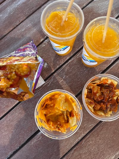 The image shows a wooden table with various snacks and beverages. Two clear cups filled with an orange beverage, possibly an orange juice or a smoothie, are on the table. One cup has a blue and yellow sticker with "Cantarito" written on it. Both cups have straws inserted in them. A clear plastic container with a yellow lid holds a portion of nacho chips topped with a red sauce and cheese. A similar container is filled with a mixture of cheese and nacho chips, possibly a cheese dip. A purple and yellow foil bag containing various toppings is also placed on the table. The image was taken in Ciudad Santa Catarina, Mexico, at coordinates 25.658 latitude, -100.438 longitude.