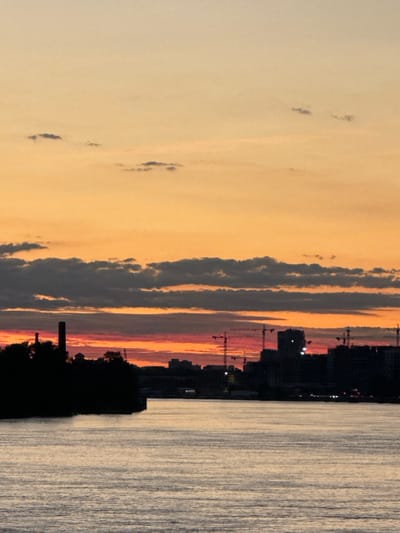 This is a photograph taken at dusk in Imeni Morozova, Russia, featuring a wide river in the foreground and a silhouetted cityscape on the horizon. The sky is painted with a gradient of warm colors, transitioning from a pale orange at the top to deeper reds and pinks near the horizon, with dark gray clouds scattered across the sky.
On the left side of the cityscape, a large, dark industrial chimney stands prominently against the sky, accompanied by silhouetted trees and buildings. Further to the right, the skyline is dominated by the silhouettes of numerous construction cranes and various buildings, suggesting ongoing development. The river in the foreground reflects the colors of the sky and appears calm with gentle ripples. The lighting conditions indicate it is either sunrise or sunset, with the sun below the horizon casting a warm glow. There are no people or discernible actions visible in the image.